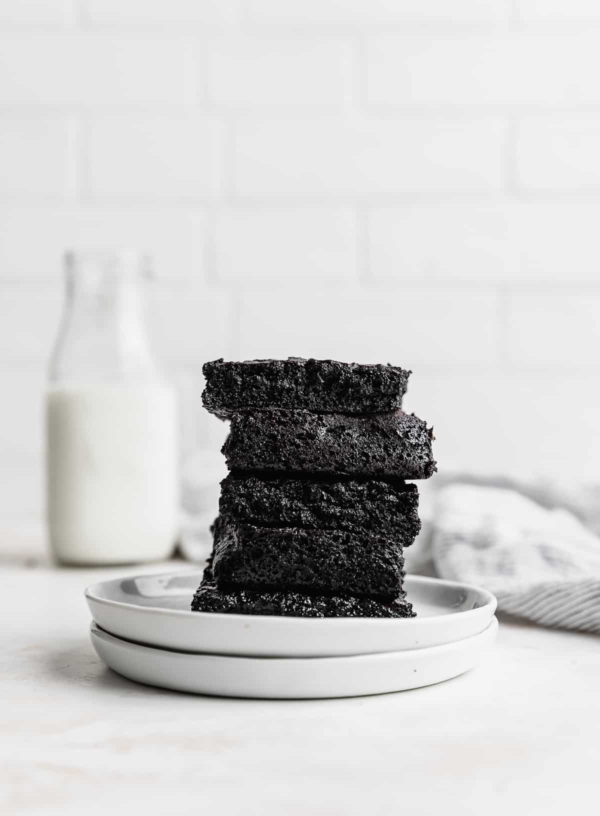 Five black cocoa brownie squares stacked on top of each other, on two white plates against a white brick background.