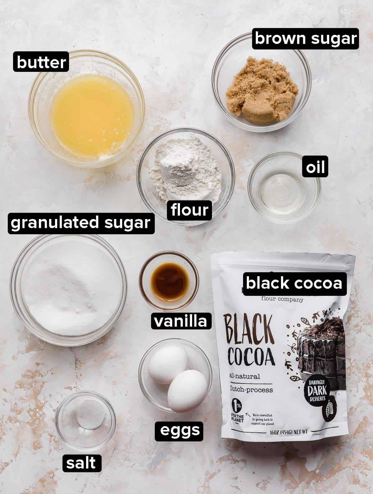 Black Cocoa Brownie ingredients portioned into glass bowls on a white textured background.