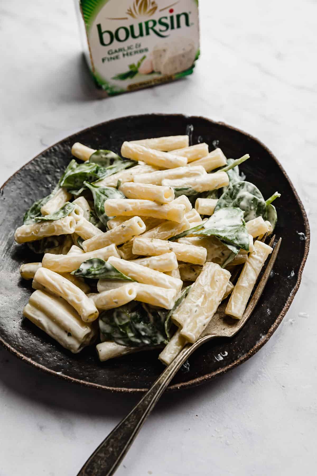 Boursin Pasta with chopped spinach on a black plate with a Boursin white and green cheese box in the background.