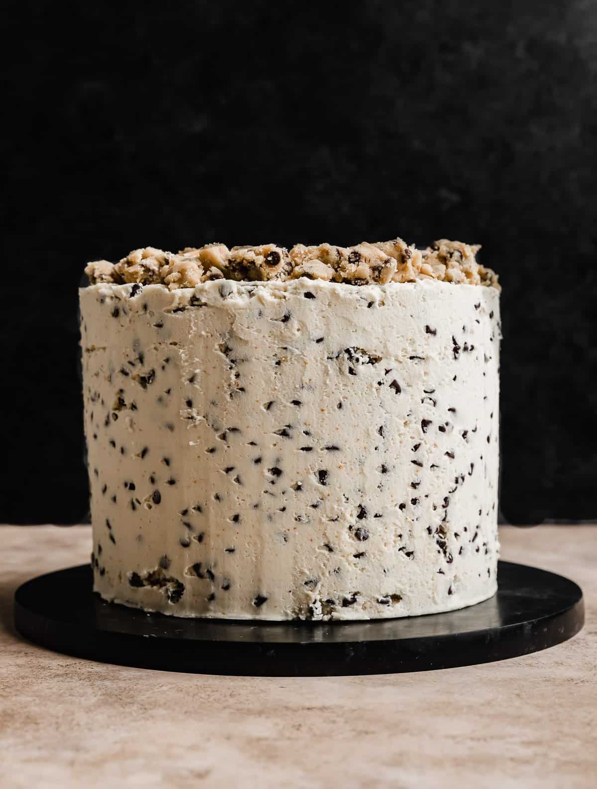 A three layered round Cookie Dough Cake frosted with white frosting that has mini chocolate chips in it, against a black background.