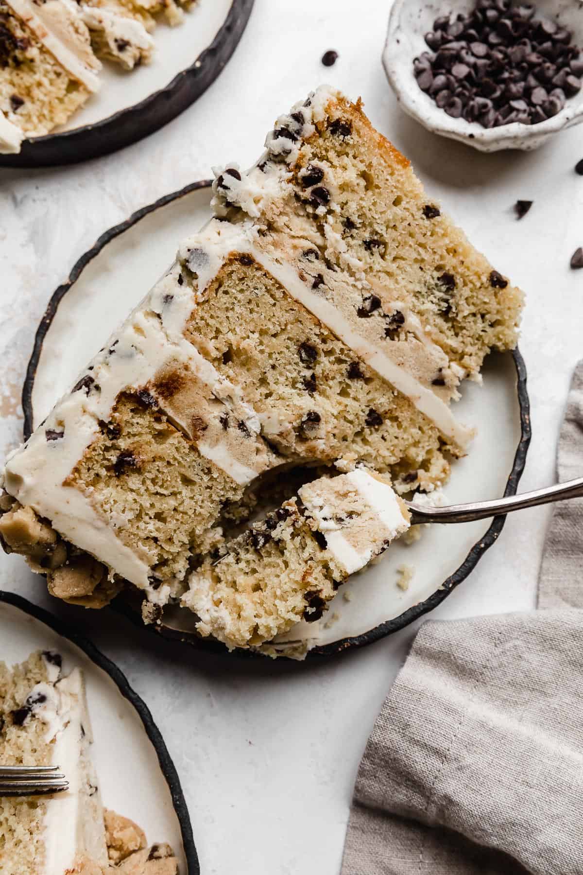 A Cookie Dough Cake slice on a black rimmed white plate with a fork cutting into the top portion of the cake.