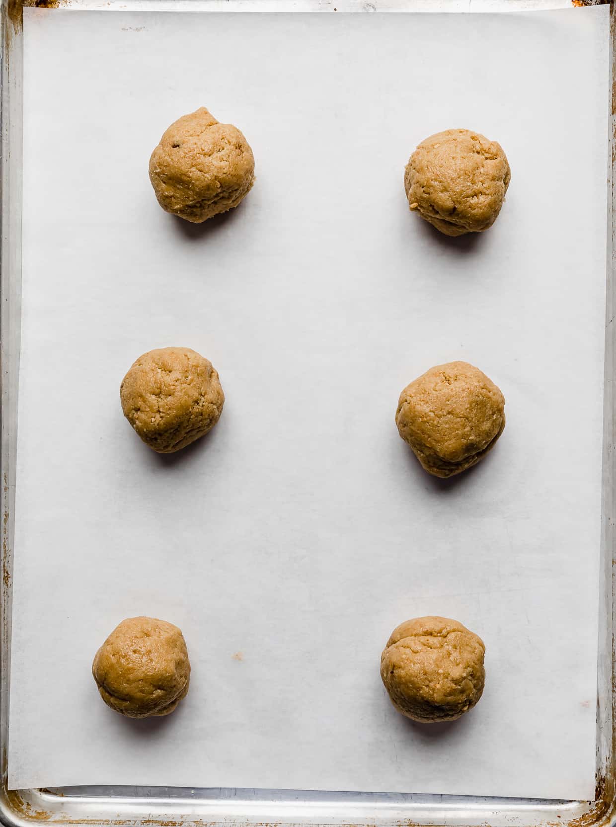 Six tan cookie dough balls on a white parchment lined baking sheet.