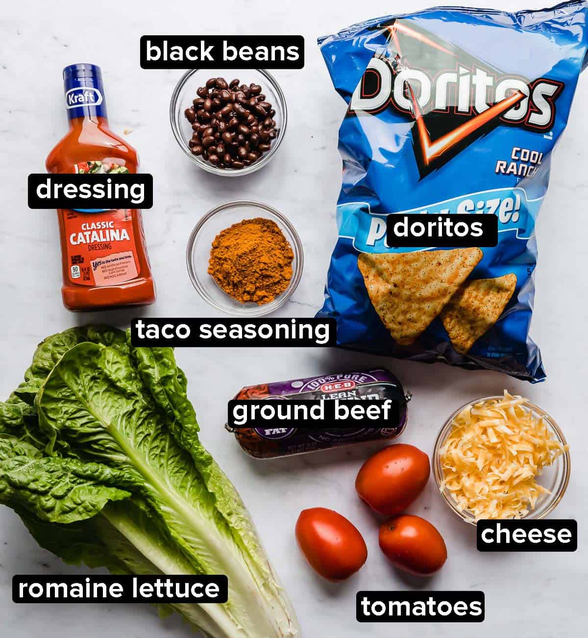 Doritos Taco Salad ingredients on a white background: ground beef, tomatoes, cheese, cool ranch Doritos, lettuce, Catalina dressing.