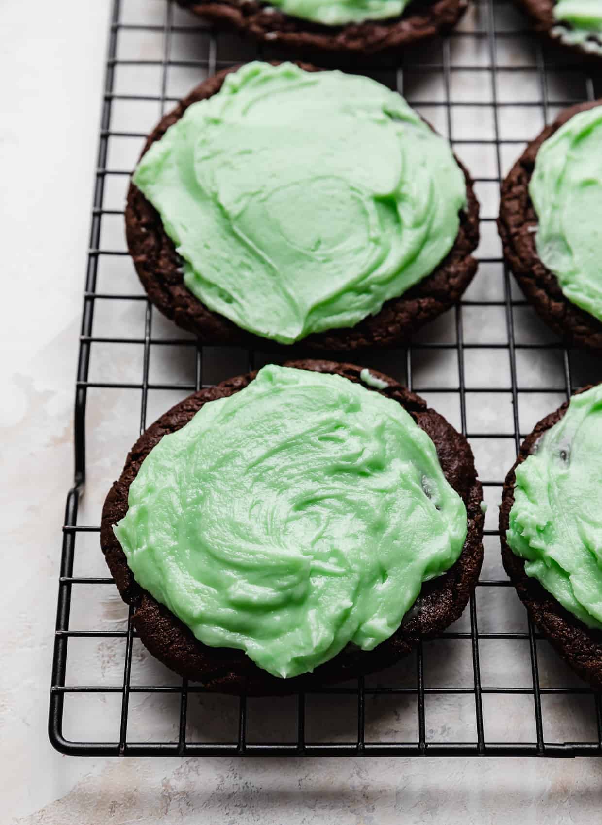 A mint colored frosting spread on a chocolate cookie that's on a wire cooling rack.