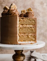 A three layer Scotcheroo Cake on a white marble cake stand against a light brown textured background.