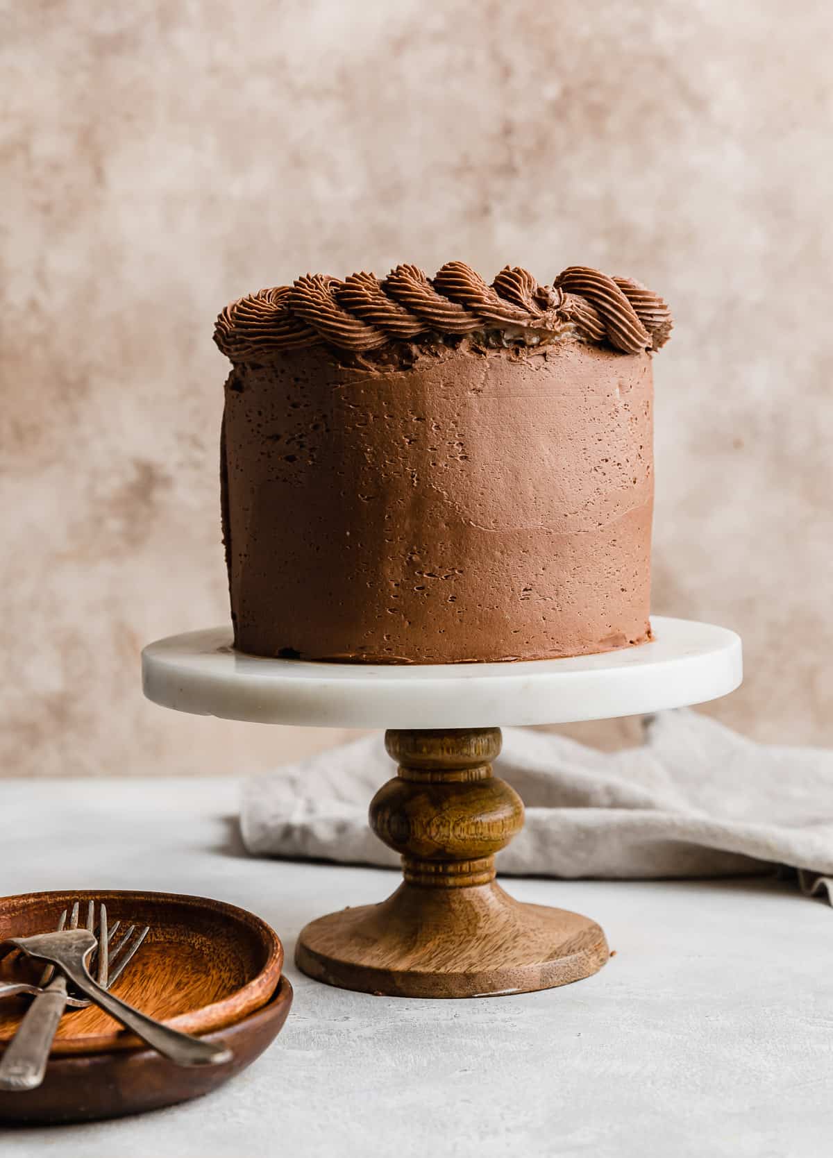 Chocolate buttercream frosting covering a small three layer German Chocolate Cake, on a cake stand against a light brown textured background.