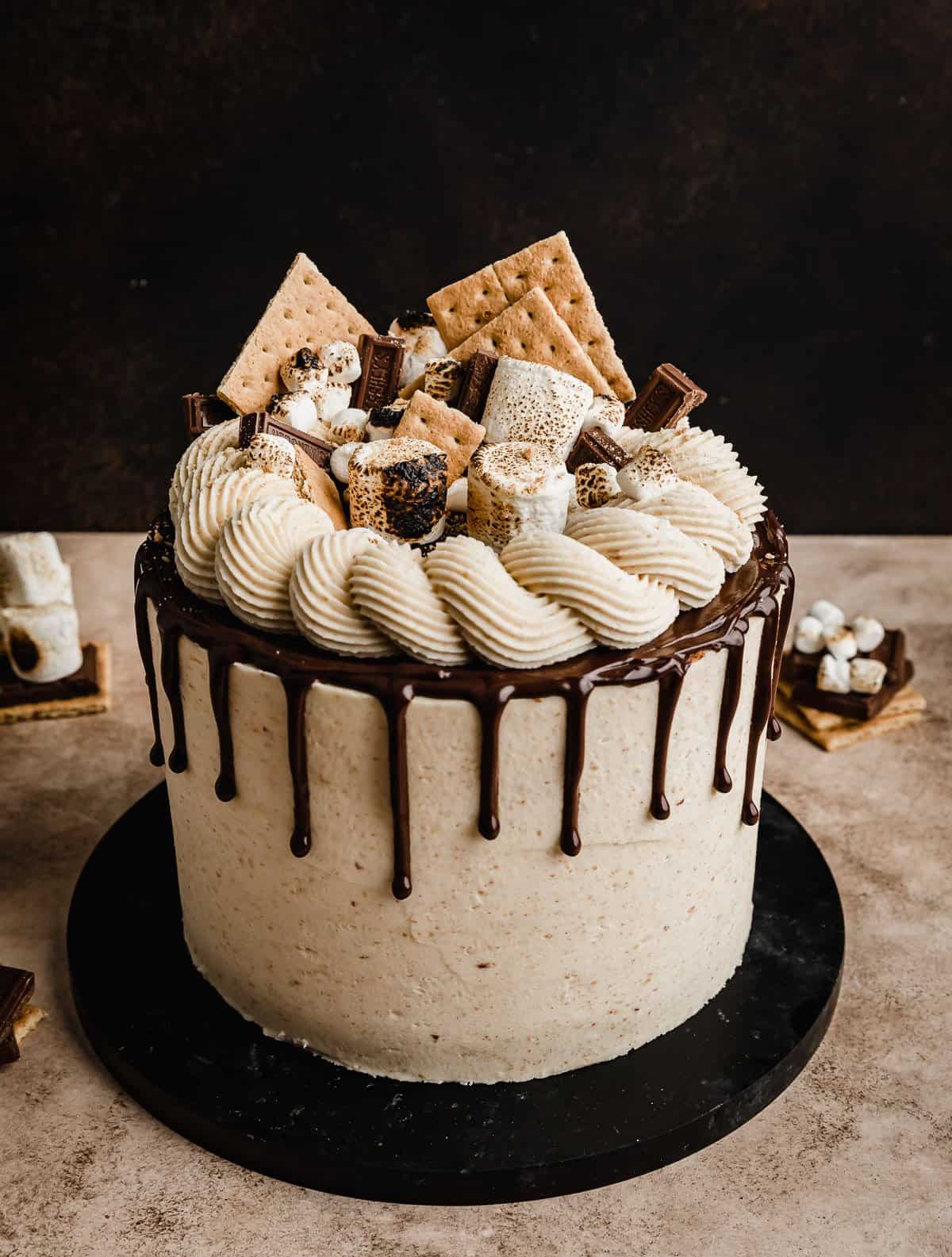 Delicious layered s'mores cake with a chocolate drip topped with graham cracker squares, toasted marshmallows and chocolate squares.