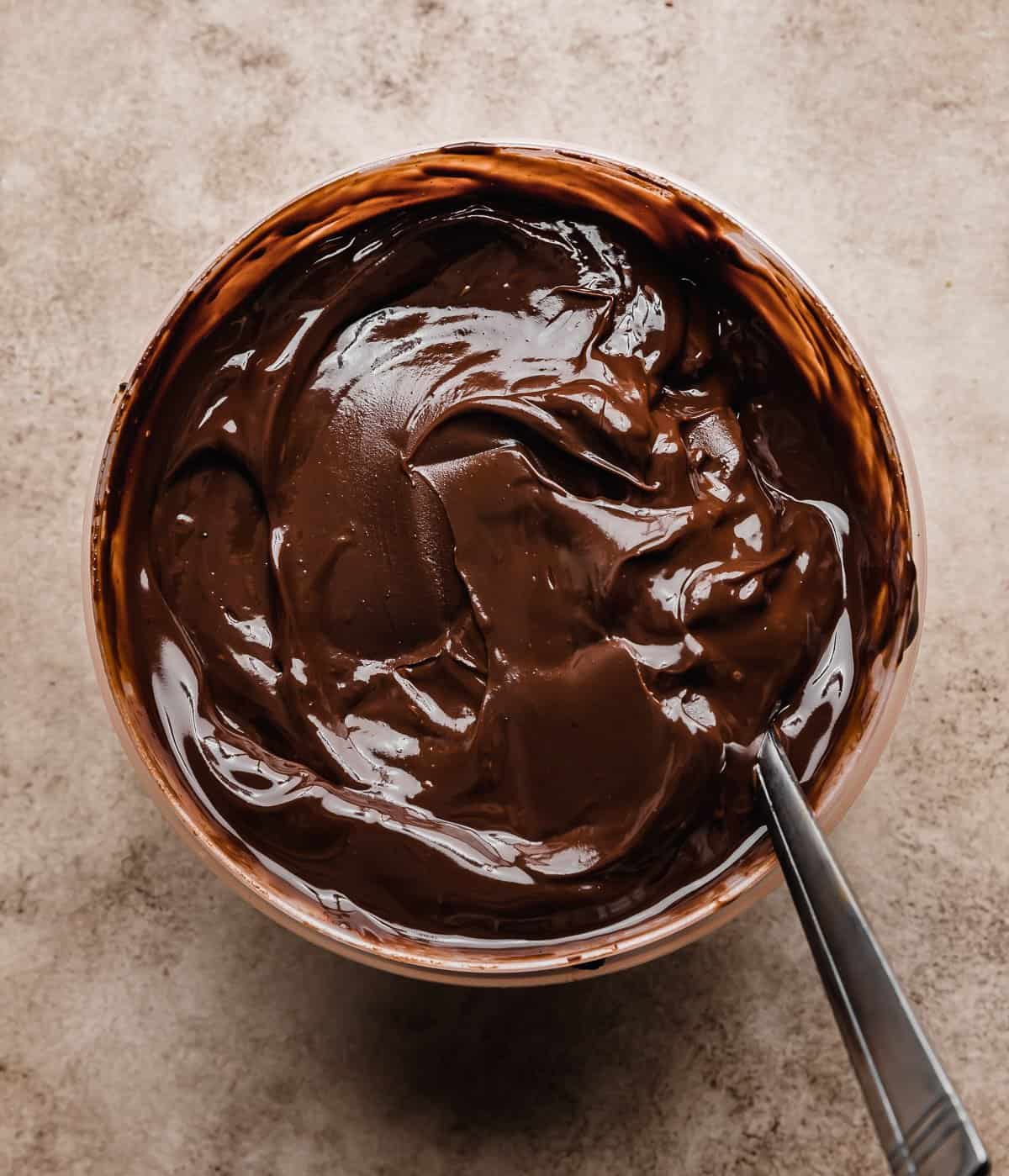 Chocolate ganache in a bowl on a beige background, to be used as a filling for a s'mores layer cake.