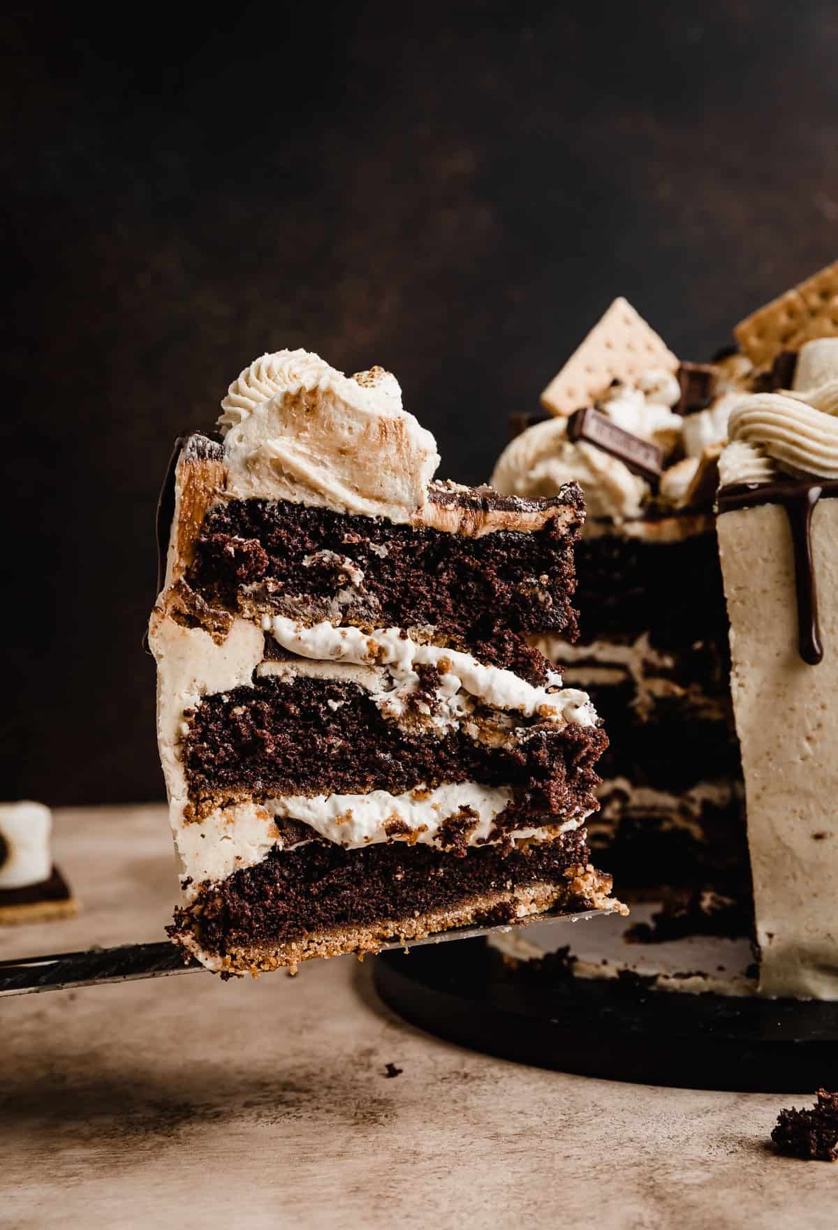 A slice of a S'mores three layer Cake balancing on a serving knife, in front of a brown background.