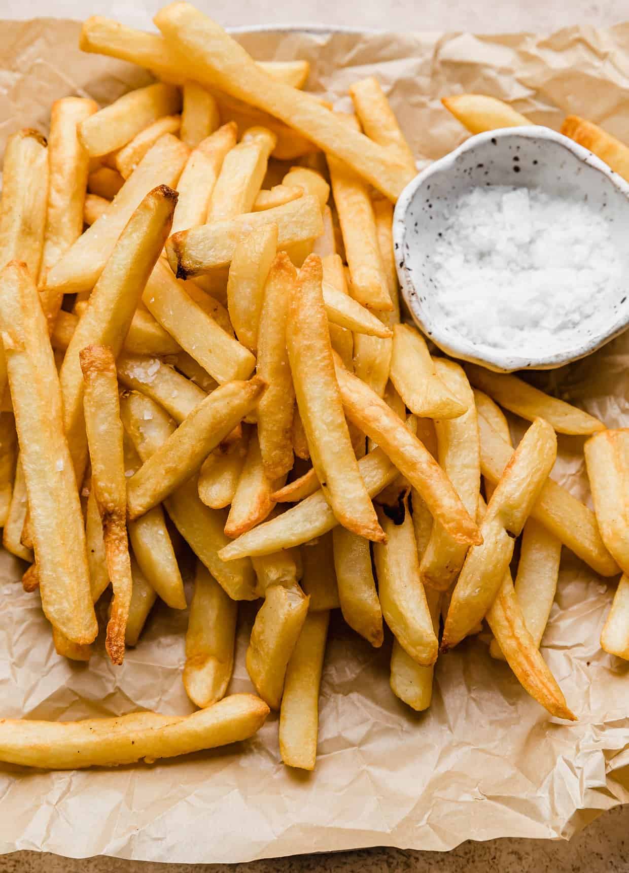 Air Fryer Frozen French Fries on a tan parchment paper with a small white bowl filled with salt.