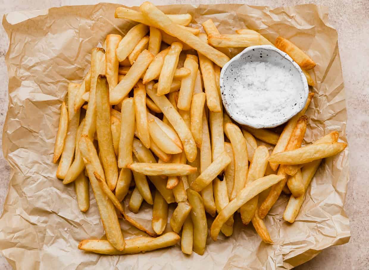 French fries made in the air fryer on a tan parchment paper next to a white bowl filled with salt. 
