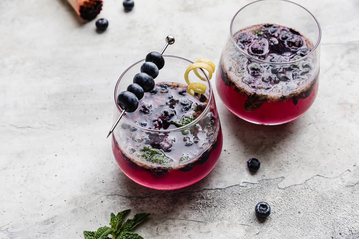 A glass cup on a gray background filled with blueberry mojito mocktail recipe.
