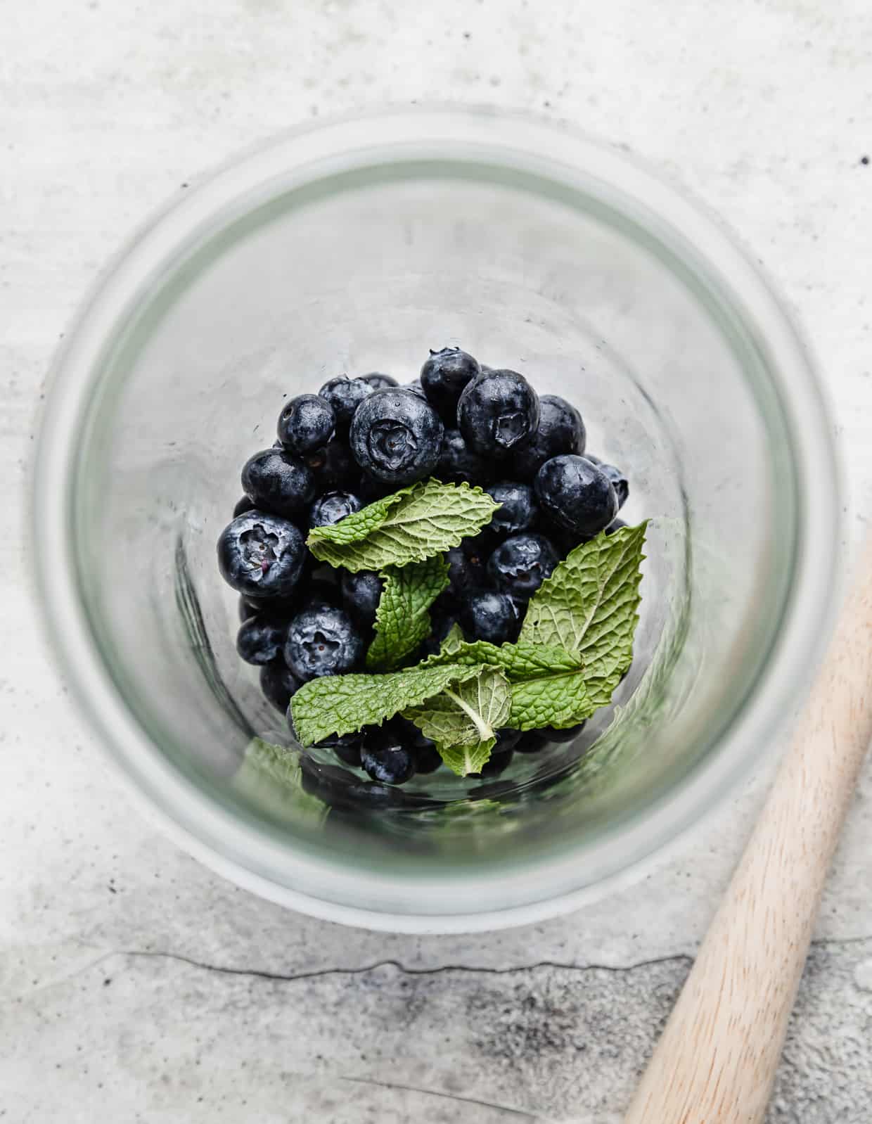 A glass jar with a handful of fresh blueberries and mint leaves in the bottom, in preparation to make a blueberry mocktail.