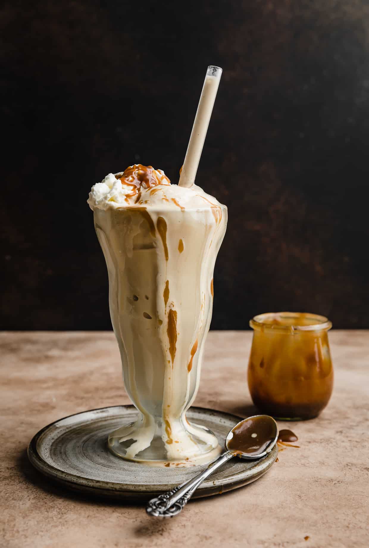 A Butterscotch Milkshake in a tall glass with a straw in it, against a brown background.