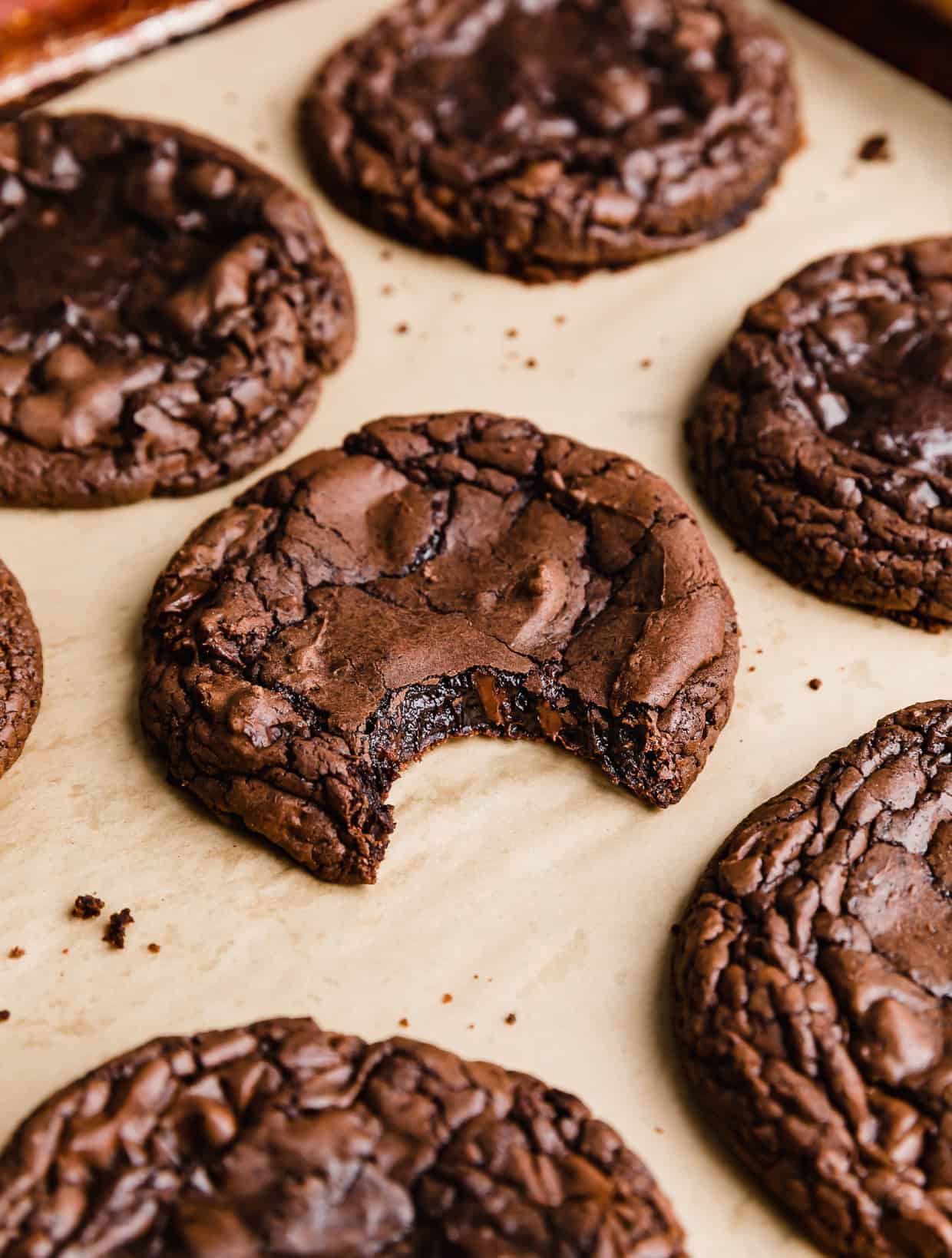 A brownie batter cookie on a tan parchment paper with a bite taken out of  the cookie.