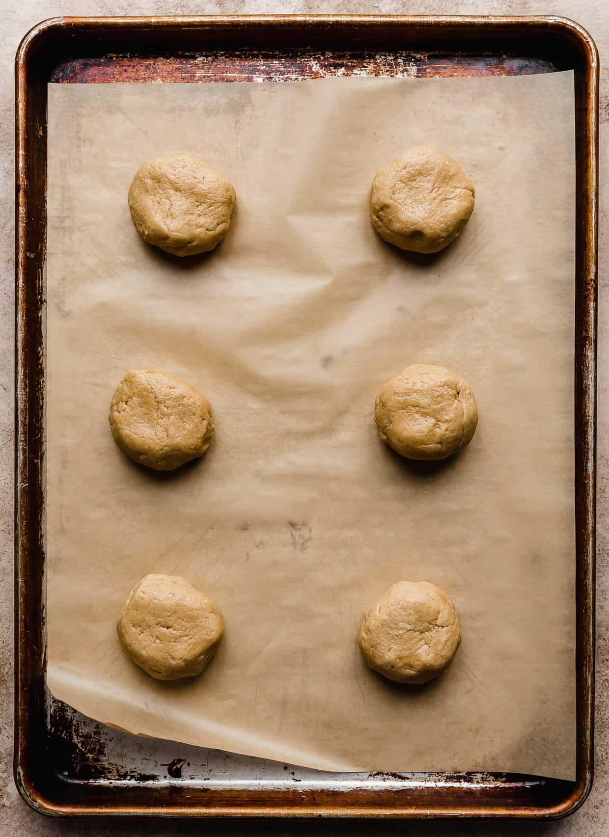 Six peanut butter cookie dough balls on a tan parchment lined baking sheet ready to make muddy buddy crumbl cookies.