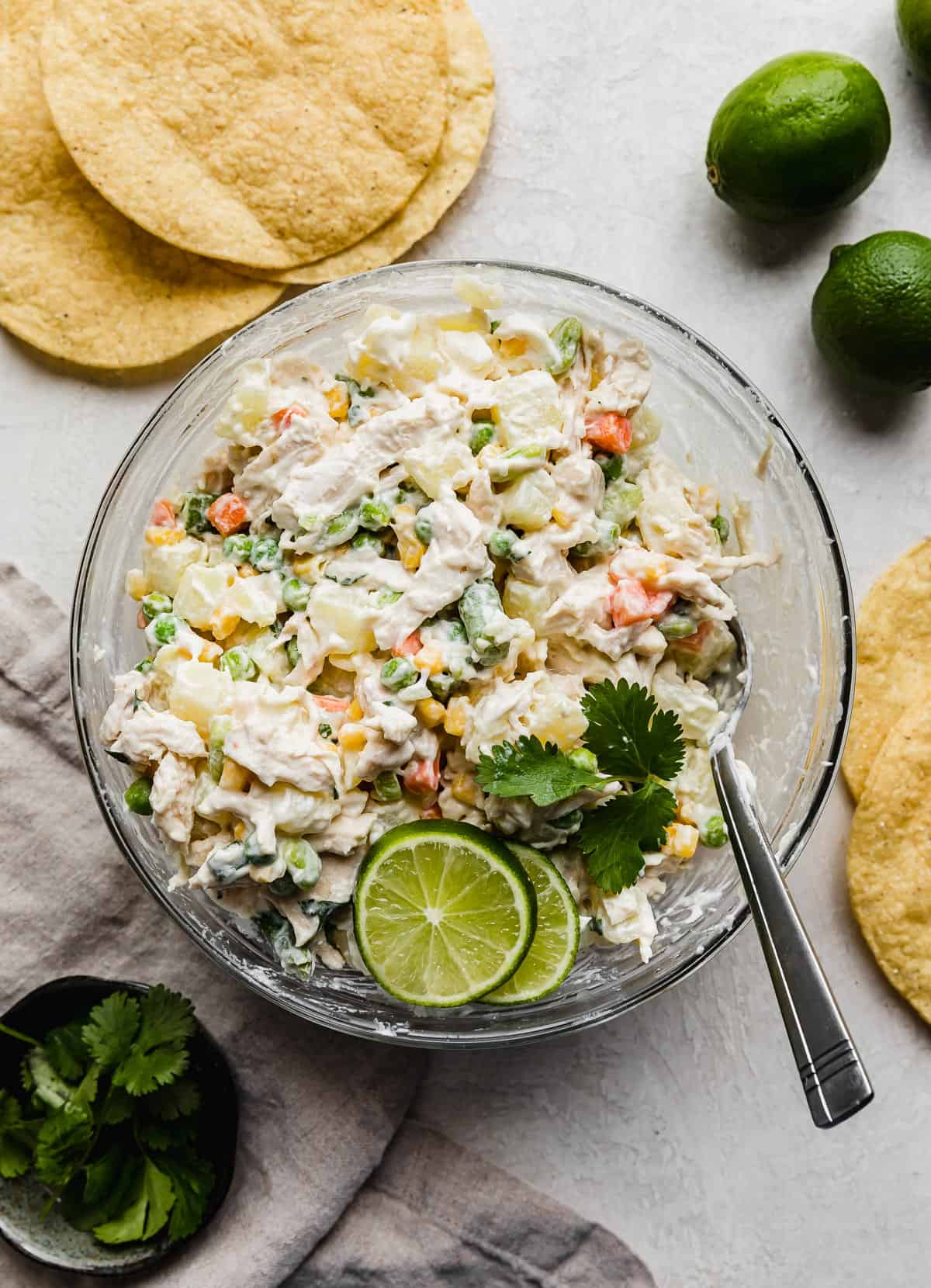 Ensalada de pollo in a glass bowl on a light grey background with tostadas and limes beside the bowl. 