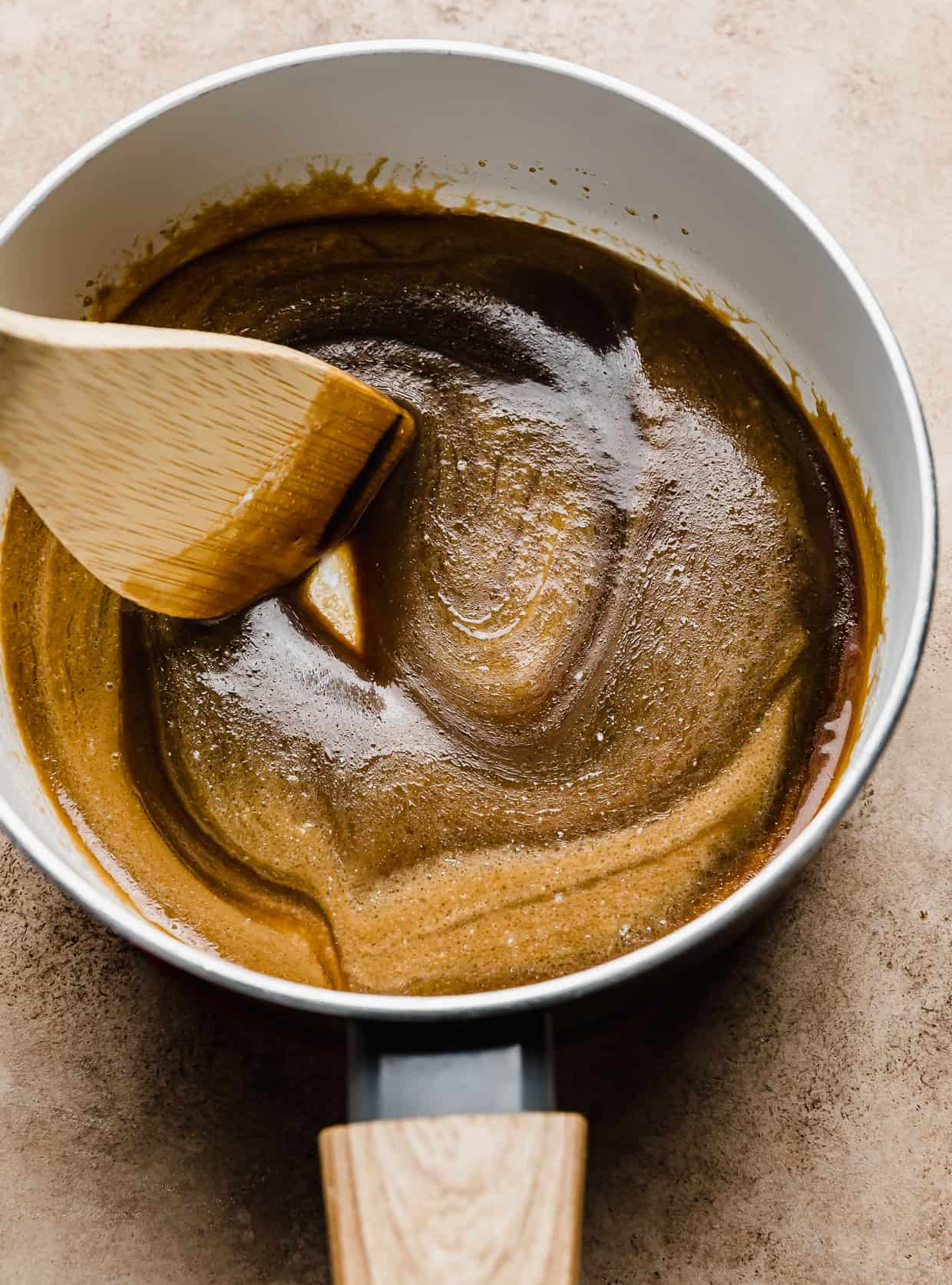 Salted Butterscotch Sauce in a white pan with a wooden spoon stirring the mixture. 