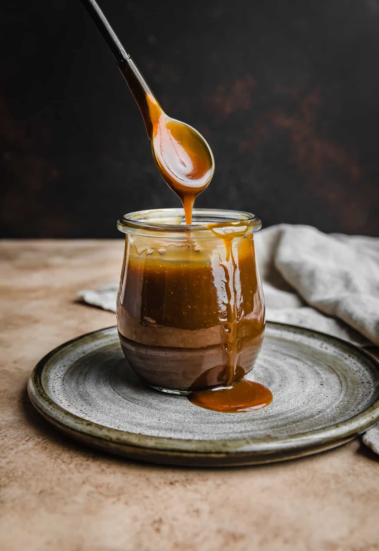A spoon lifting out Salted Butterscotch topping from a small glass jar, against a dark brown background.