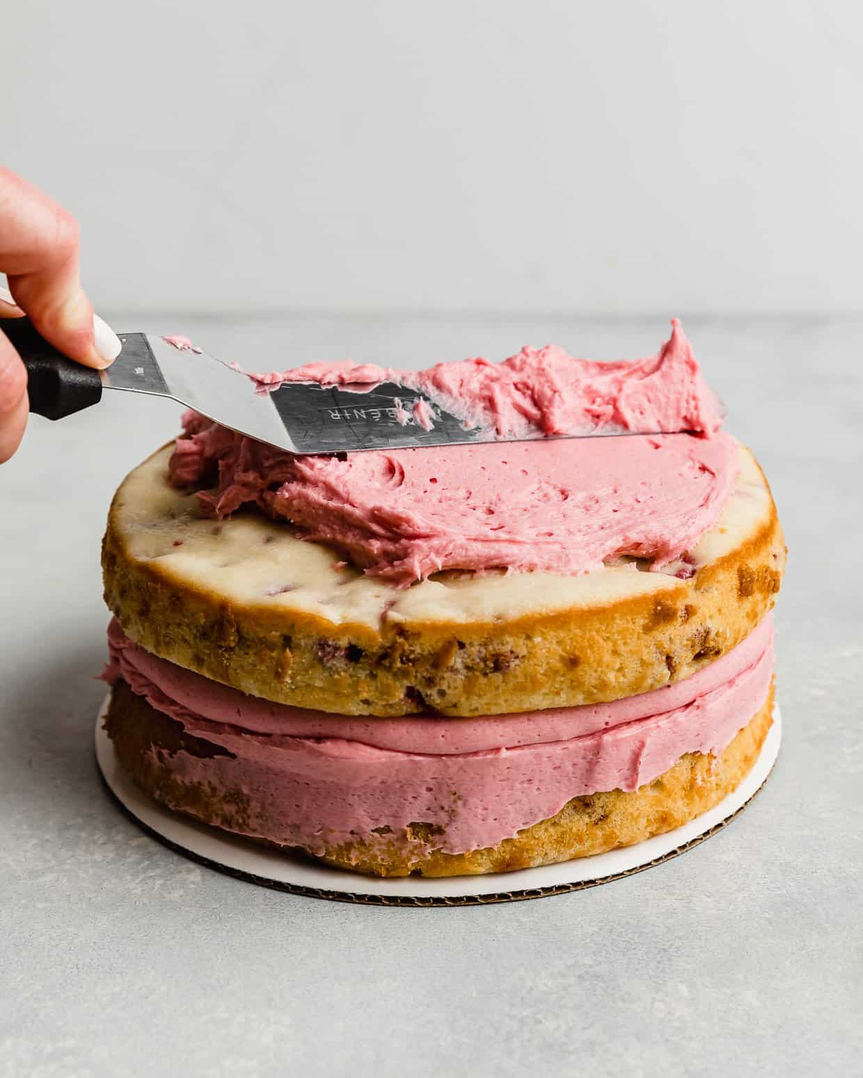 A spatula spreading a pink raspberry frosting overtop two layer cake.
