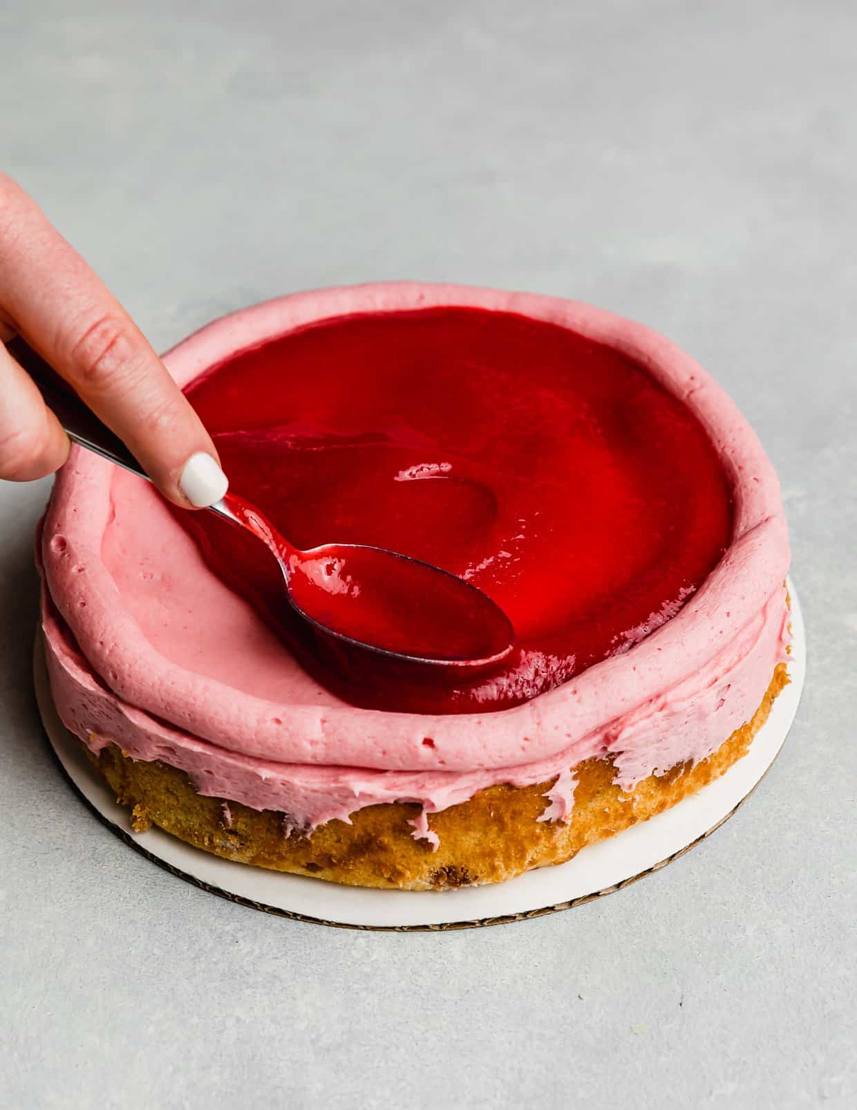 A spoon spreading a raspberry filling overtop a round cake layer that has a buttercream dam piped around the perimeter.
