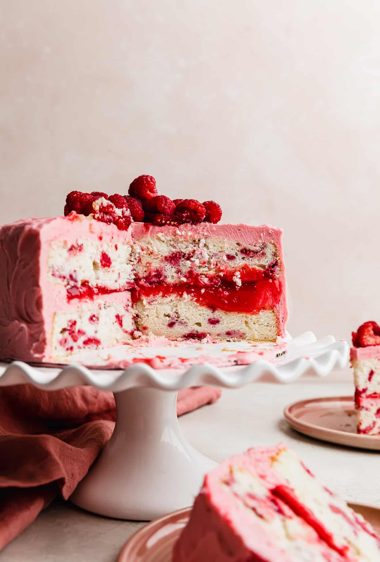A two layer Lemon Raspberry Cake on a white cake stand with a slice of cake removed showing the raspberry filling between each layer.