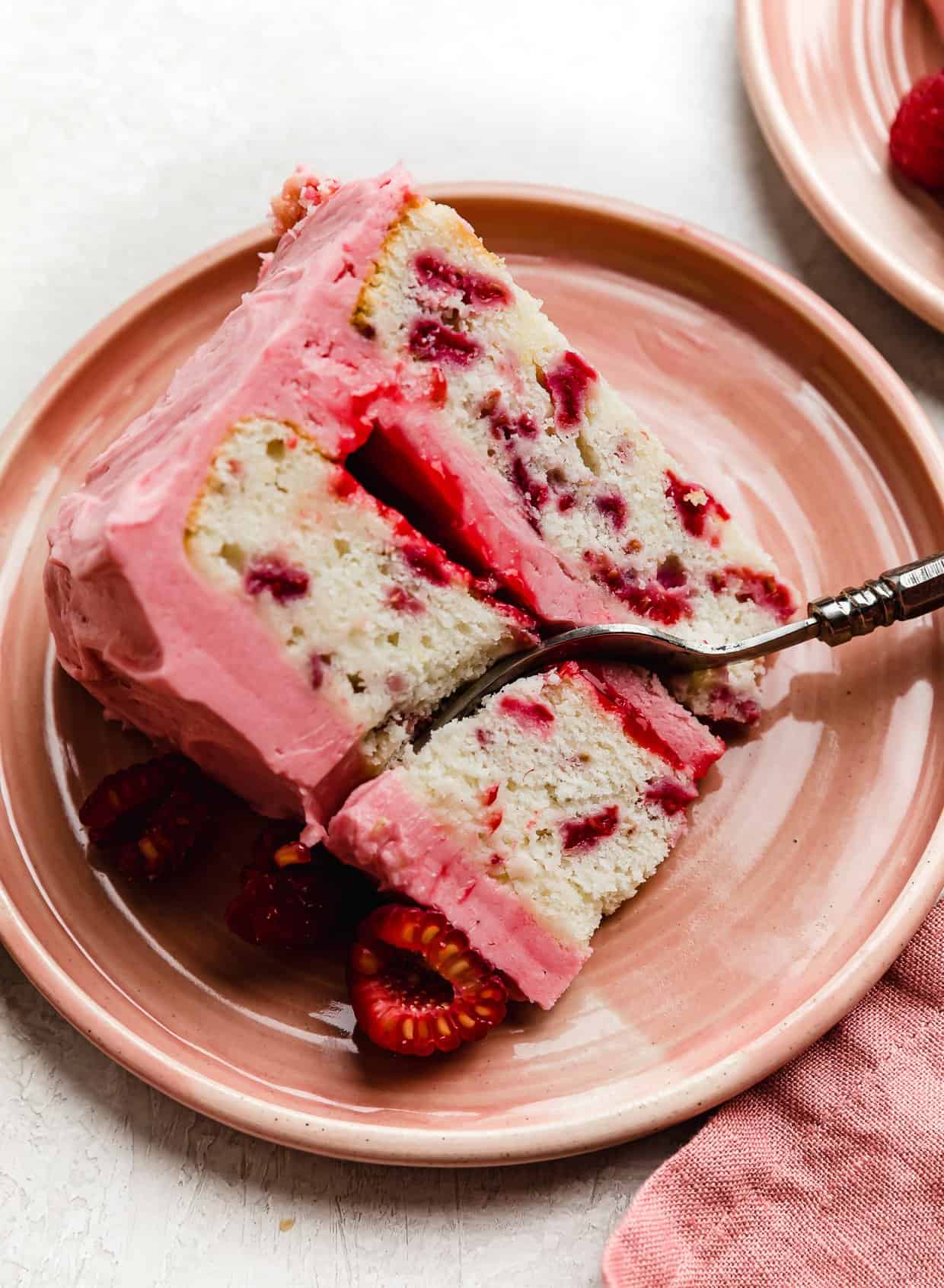 A slice of Lemon Raspberry Cake that has raspberry filing between each layer, on a pink plate with a fork cutting into the cake.