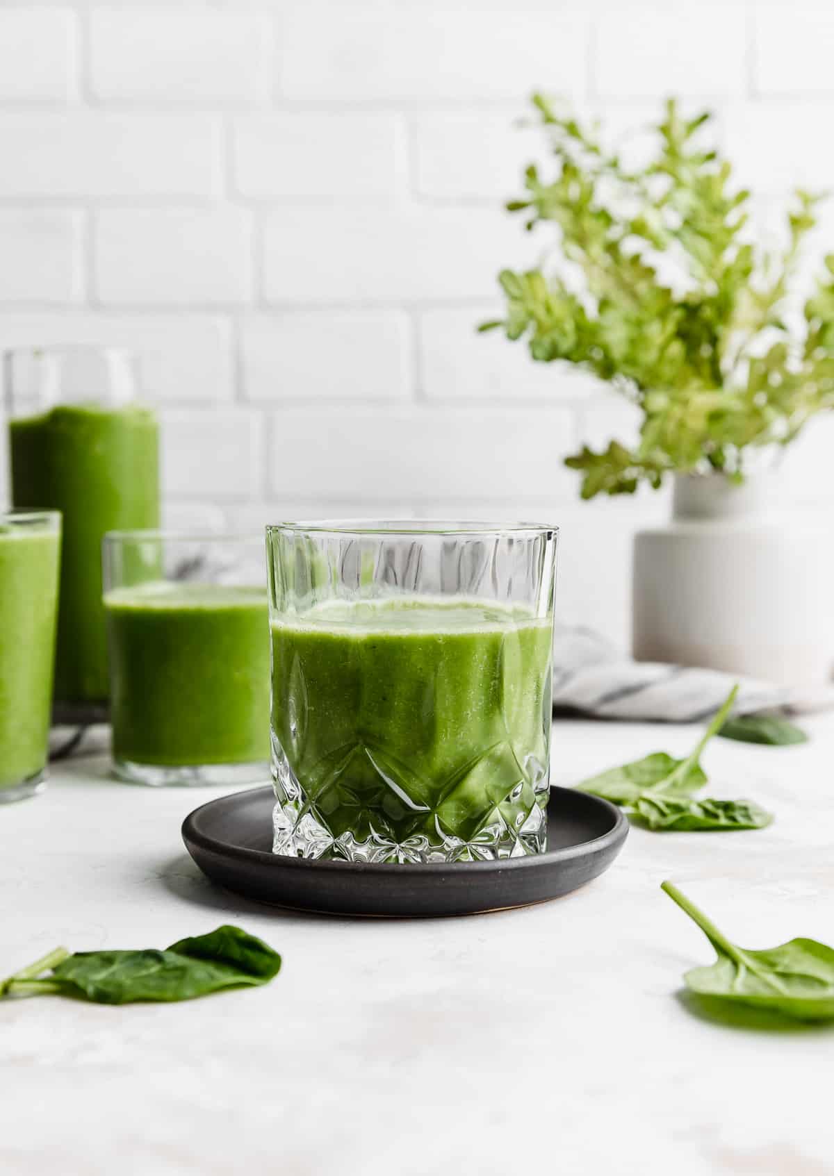 A textured clear glass on a black plate that's filled with a dark green Pineapple Spinach Smoothie.