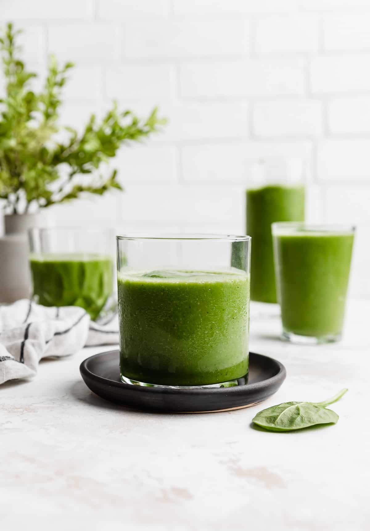 A small clear glass filled with dark green Pineapple Spinach Smoothie against a white brick background.