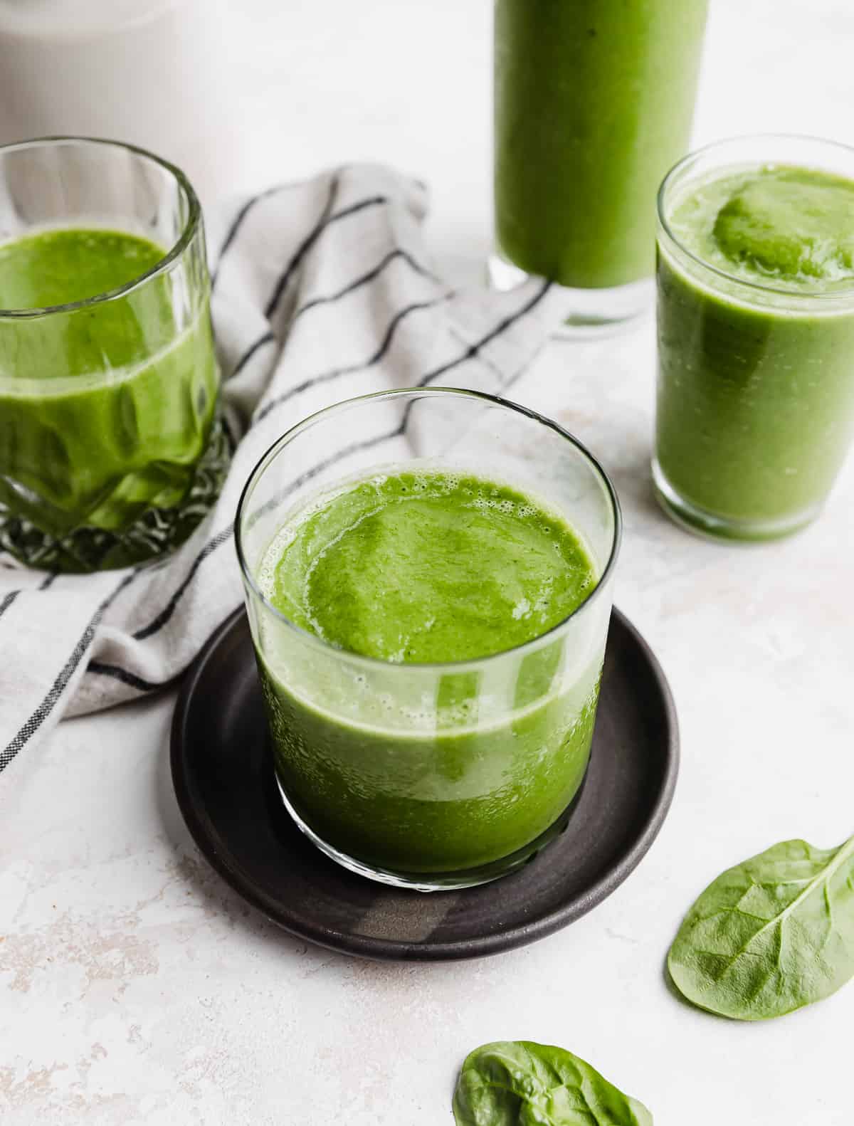 A clear glass filled with a green Pineapple Spinach Smoothie on a black plate, with three more glasses in the background that are filled with a green smoothie too.
