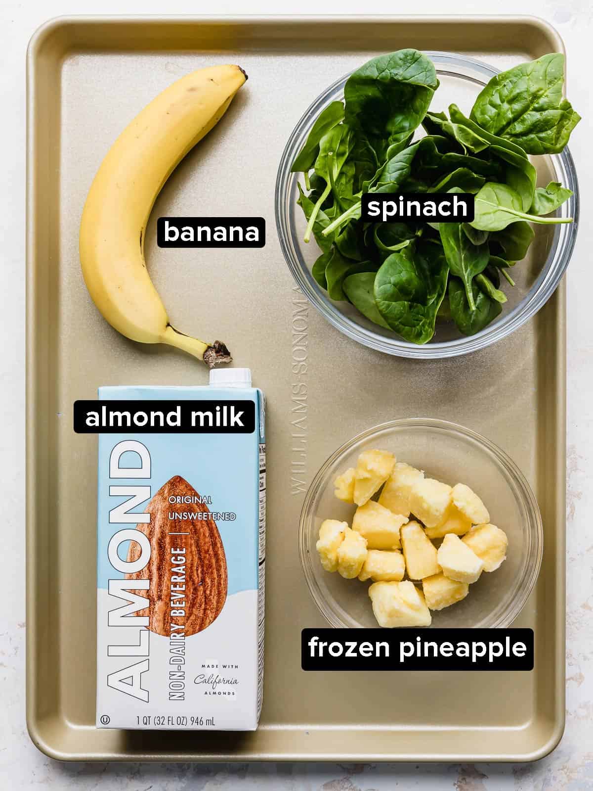 Ingredients used to make Pineapple Spinach Smoothie on a bronze baking sheet (banana, spinach, pineapple, almond milk).