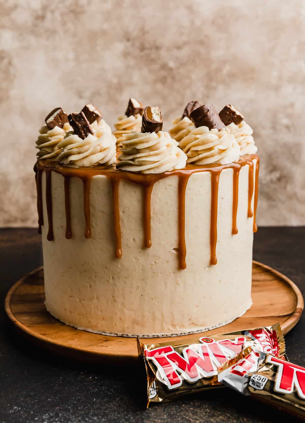 A three layer Twix cake recipe frosted with a tan shortbread buttercream frosting and a caramel drip going down the cake, topped with frosting swirls and Twix pieces.