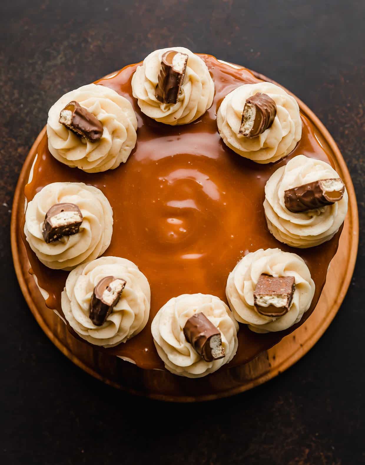 Overhead photo of a layered Twix cake topped with caramel, frosting swirls, on a brown background.
