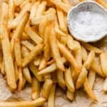 Golden French fries on a parchment paper with salt in a white bowl next to the fries.