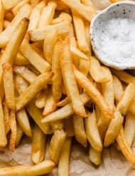 Golden French fries on a parchment paper with salt in a white bowl next to the fries.