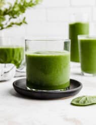 A small clear glass filled with a dark green Pineapple Spinach Smoothie, against a white brick background.