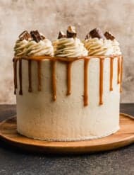 A Twix cake covered in a tan buttercream frosting, caramel drip, frosting swirls and Twix pieces.
