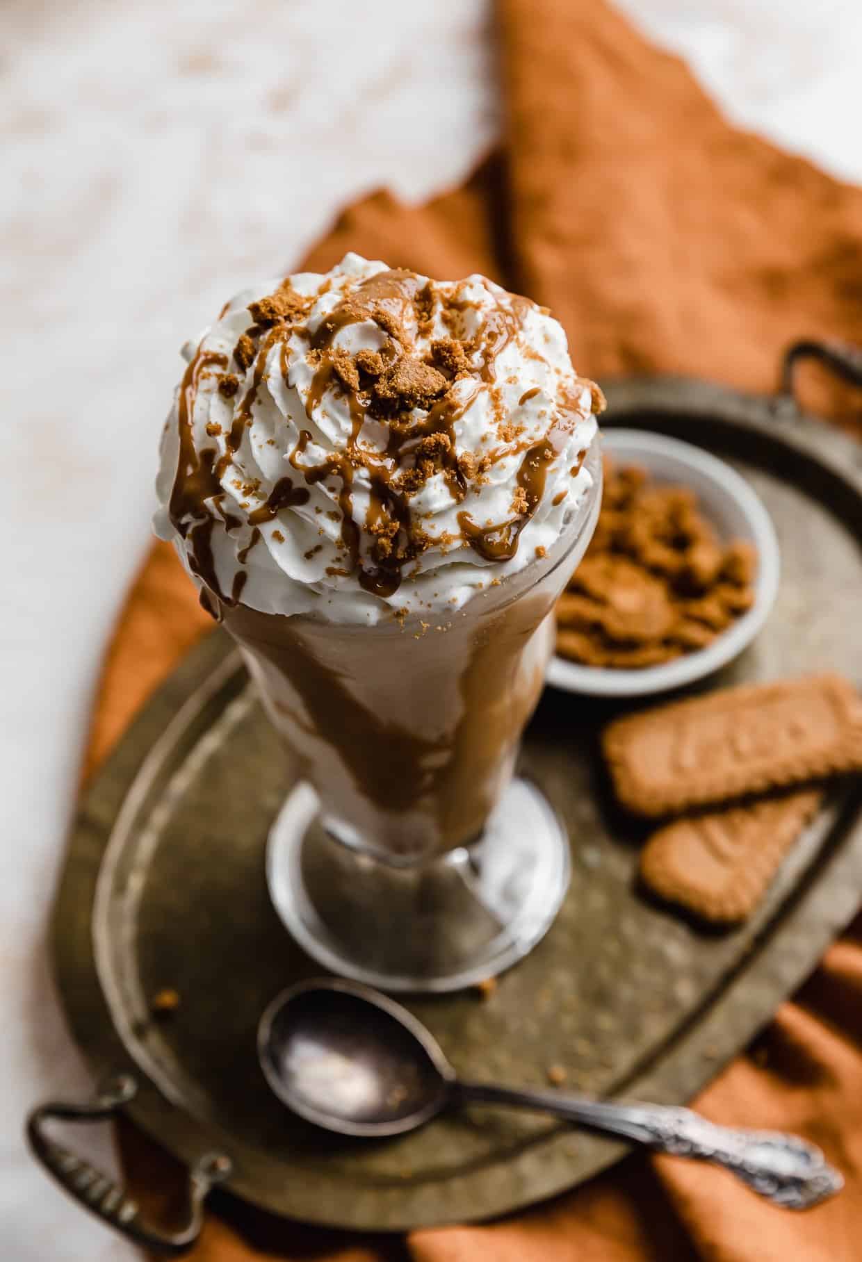 A Biscoff Milkshake in a glass cup on a bronze tray with Biscoff cookies in the background.