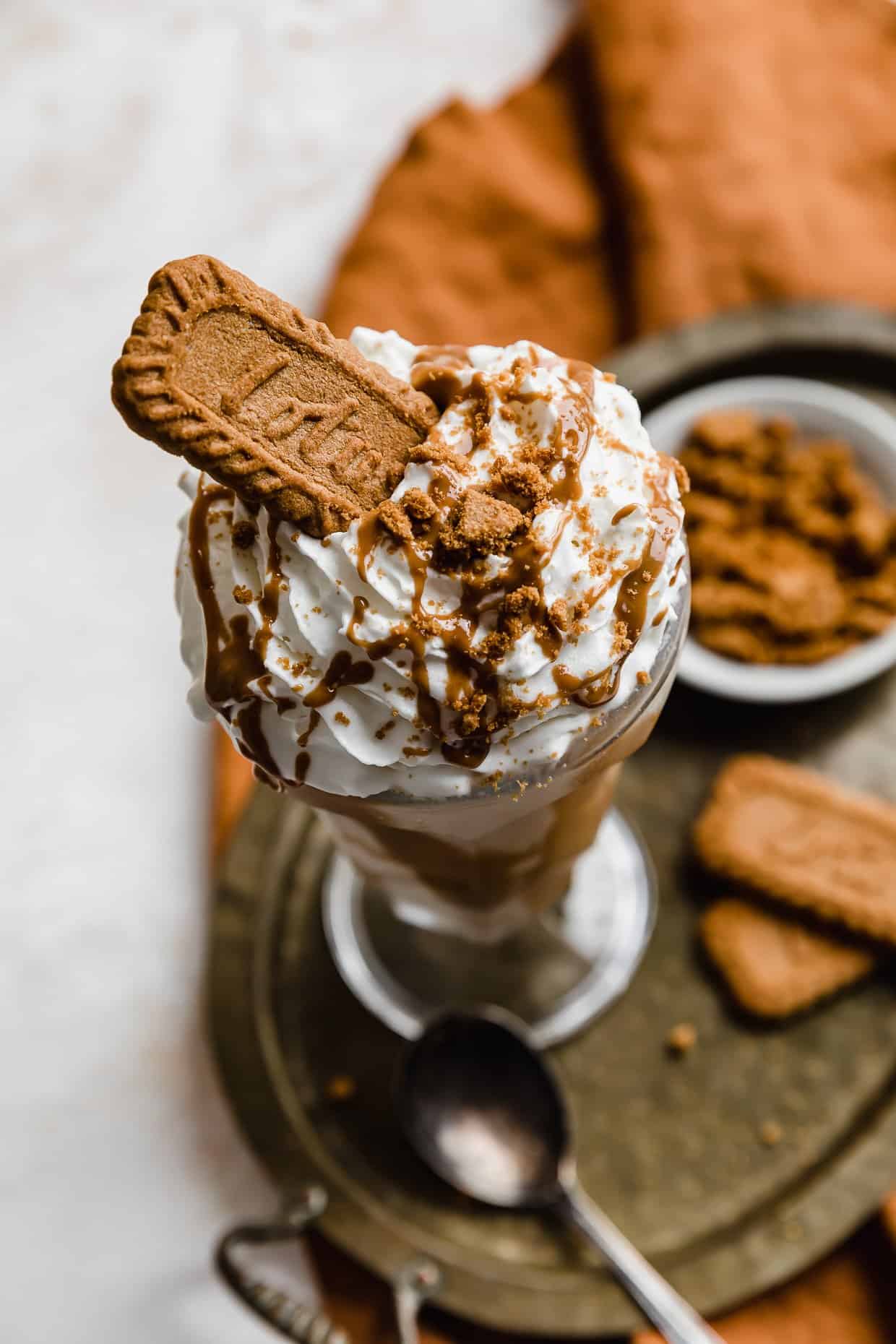 Biscoff Milkshake topped with whipped cream, biscoff drizzle and a lotus cookie, on a bronze tray that's on a white background. 