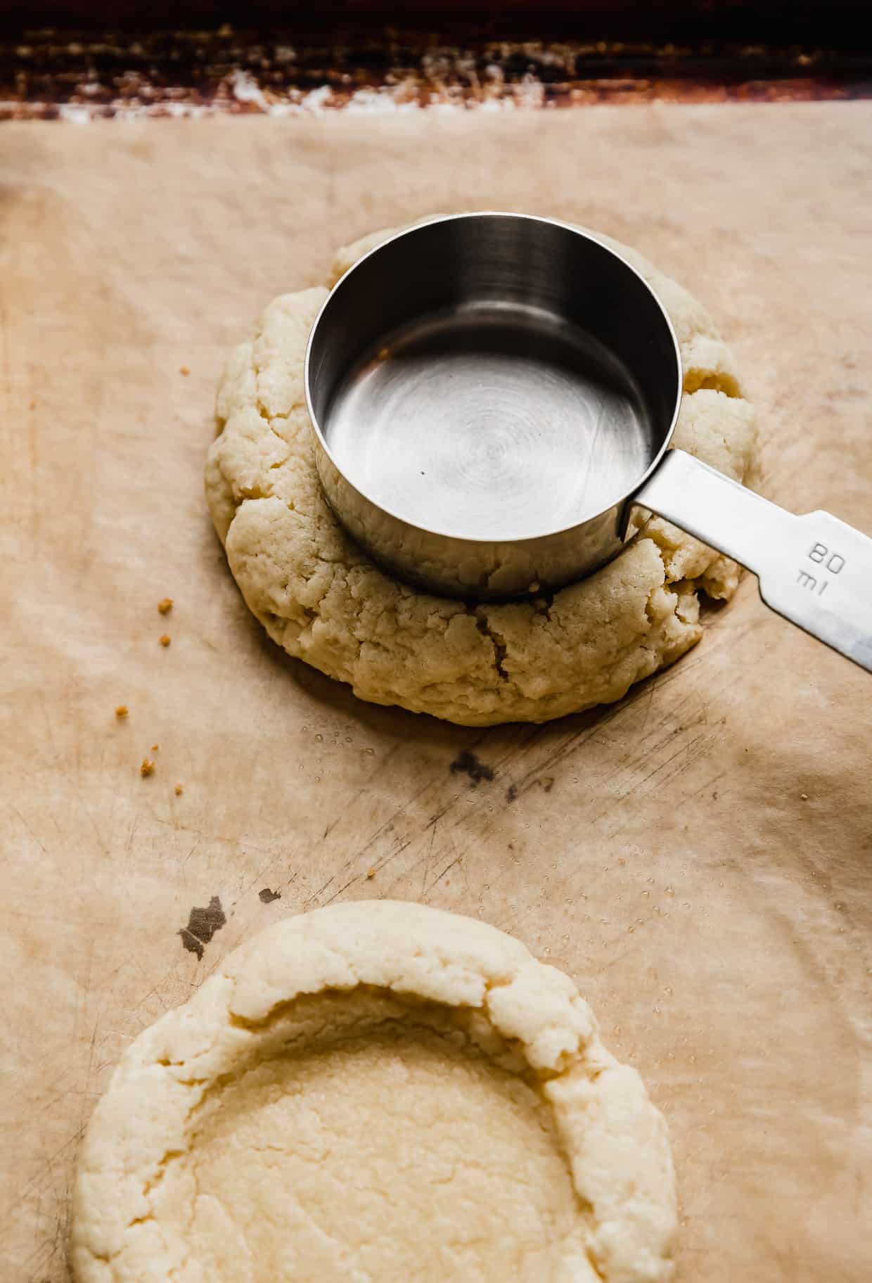 A third cup measuring cup pressing into a partially baked sugar cookie on a baking sheet.