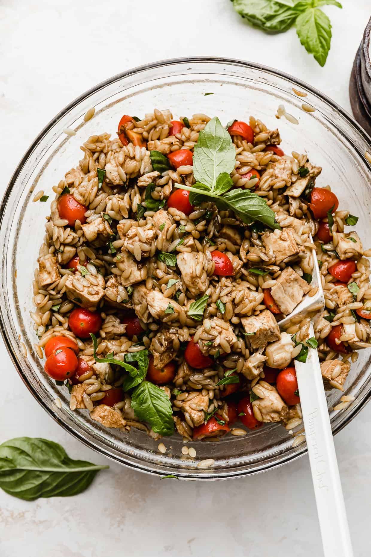 Chicken Caprese Orzo Salad in a glass bowl with a white spatula stirring the salad.