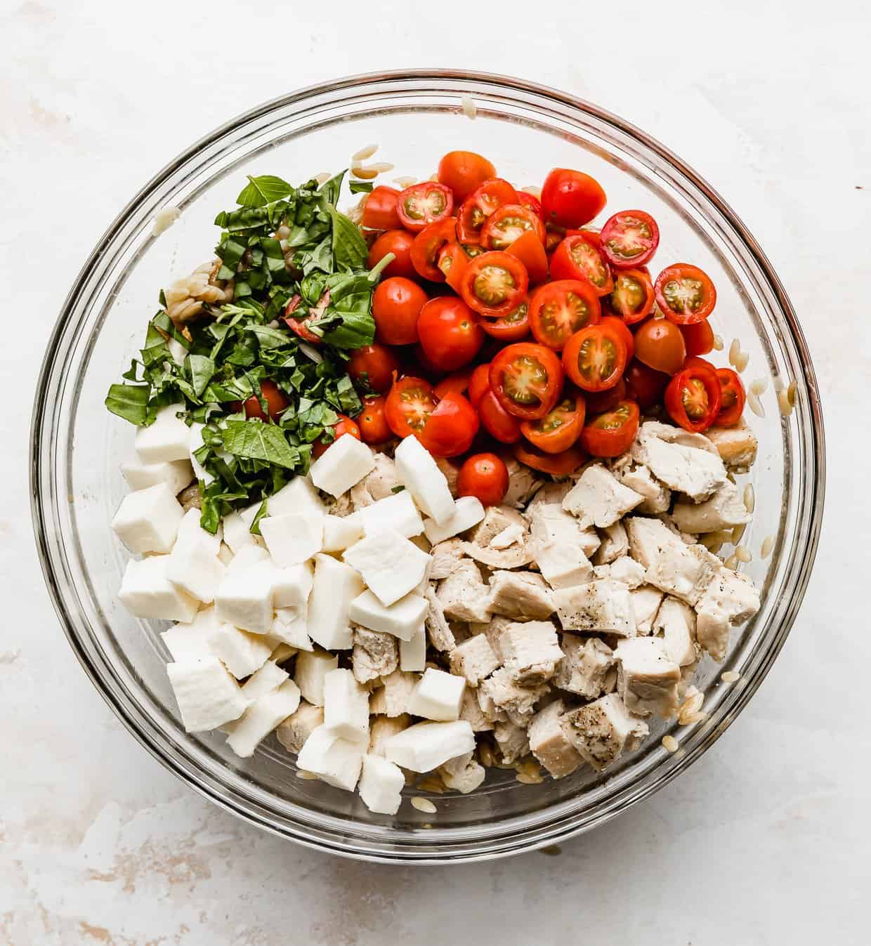 Glass bowl with orzo caprese salad ingredients in it: mozzarella, tomatoes, basil, chopped chicken, orzo pasta.