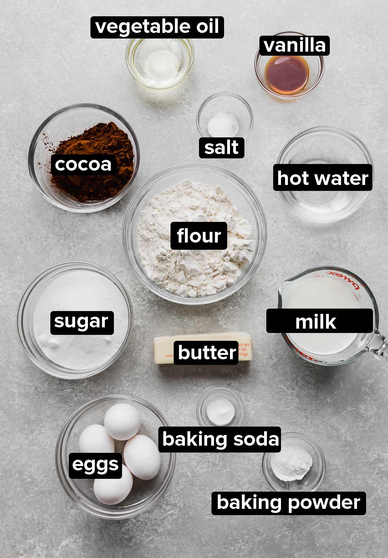 Ingredients used to make Chocolate Cake with Peanut Butter Frosting portioned into glass bowls on a light gray textured background.