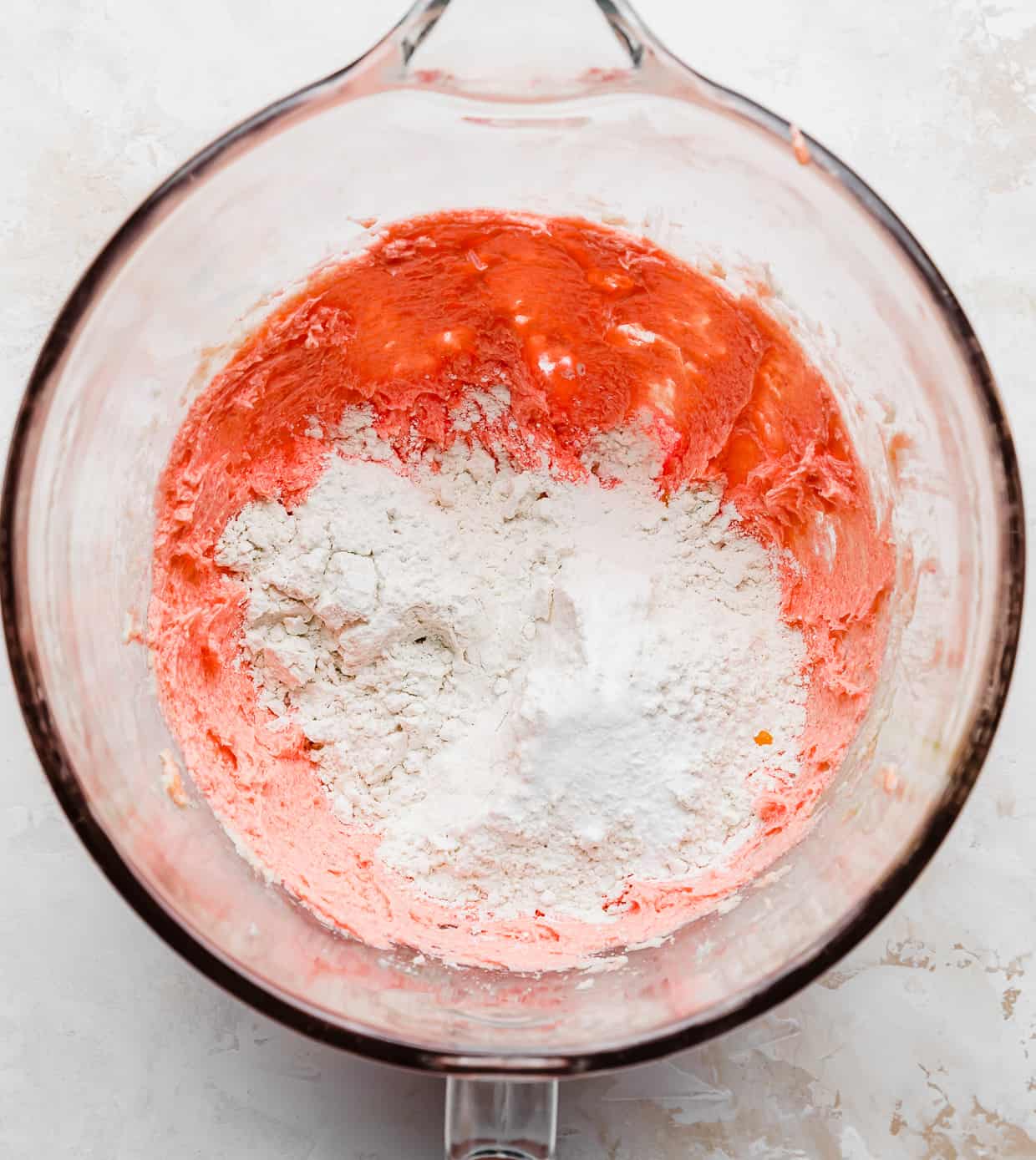 Flour overtop of pink cookie batter in a glass bowl.