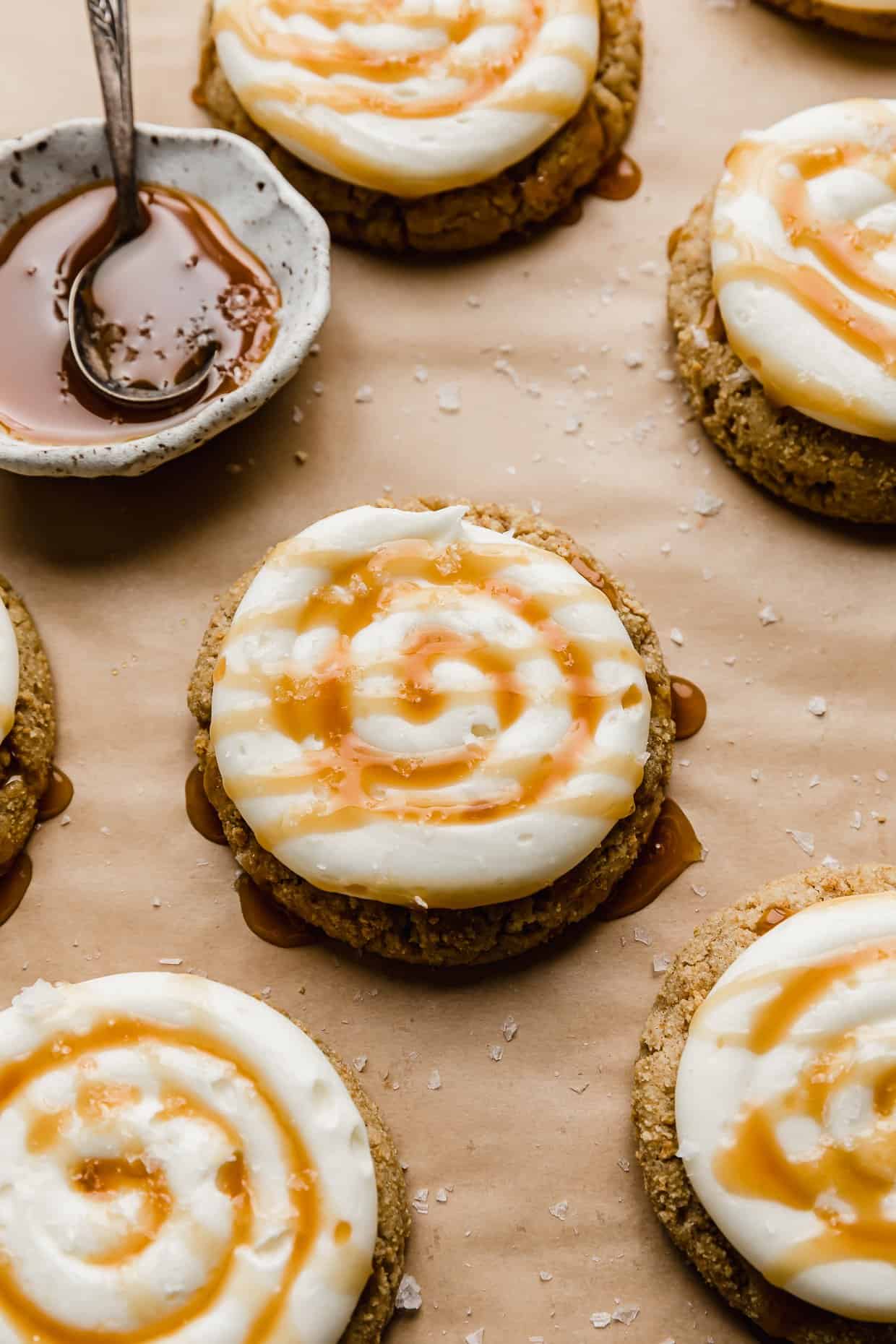Crumbl Salted Caramel Cheesecake Cookies on a tan parchment paper, each cookie topped with a swirl of caramel cream cheese frosting, caramel, and flaky sea salt.