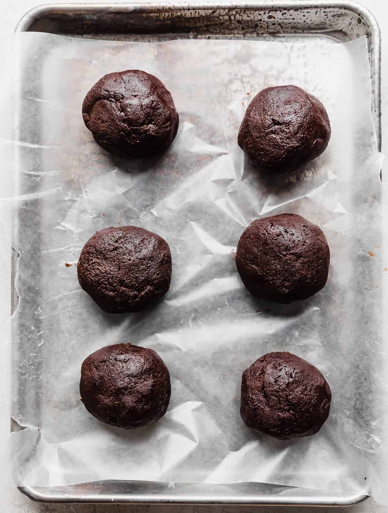 Six Crumbl Molten Lava Cookie dough balls on a wax paper lined baking sheet.