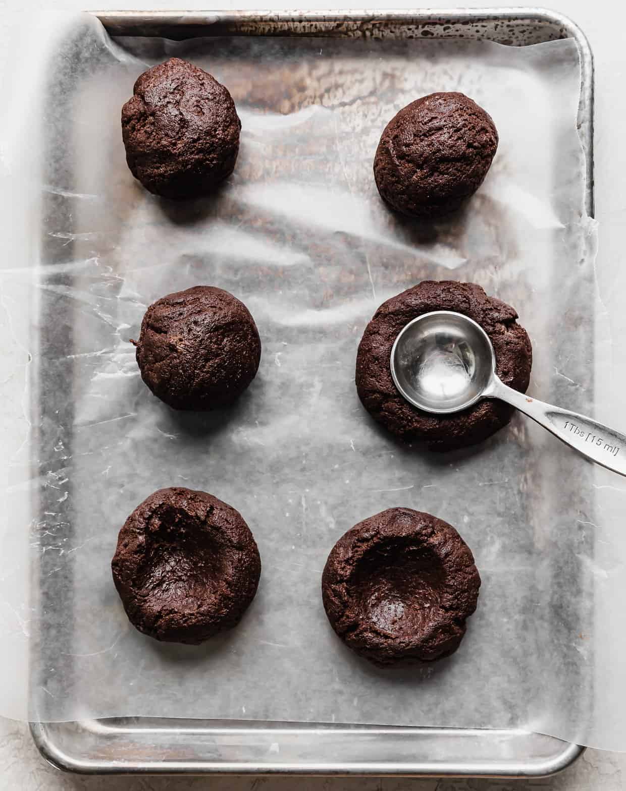 Six chocolate cookie dough balls with a metal tablespoon pressing into the center of one of the balls to create an indentation.