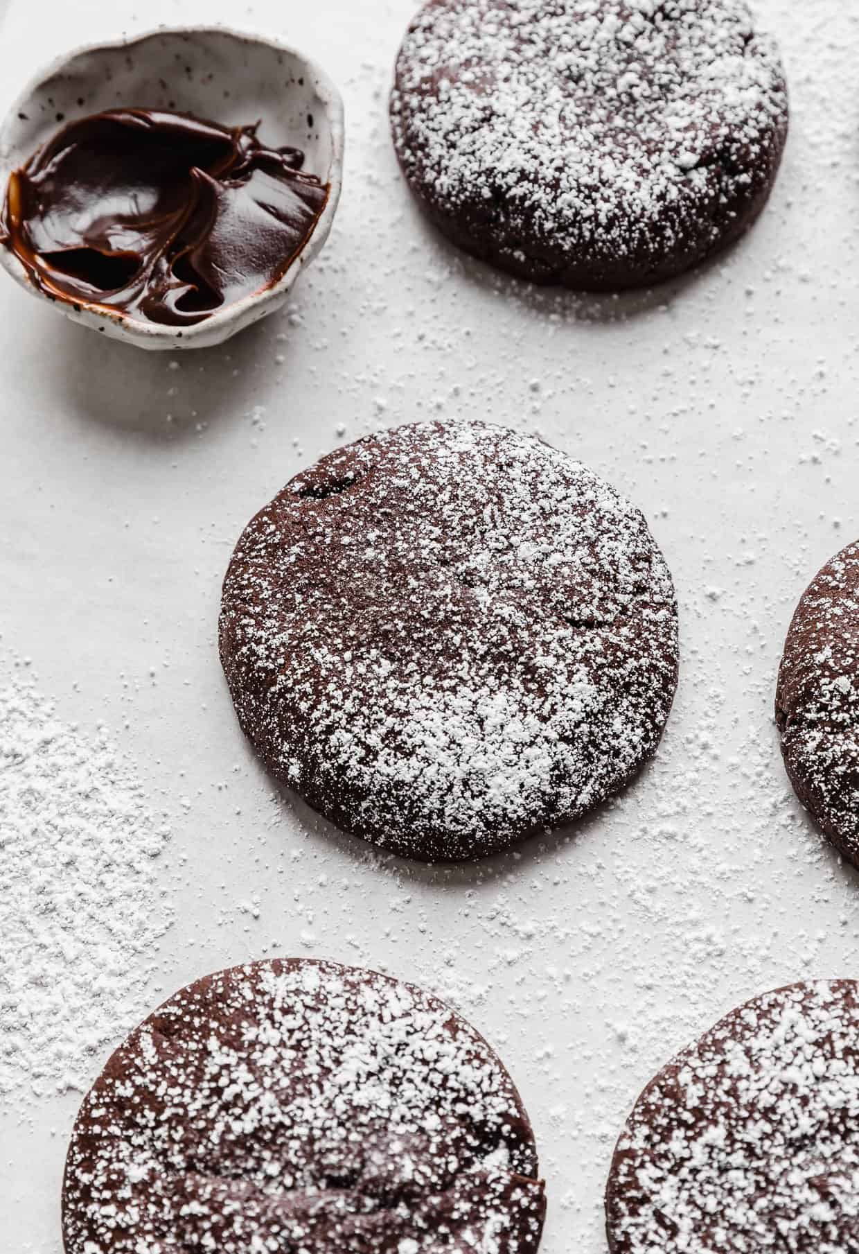 Powdered sugar dusted Crumbl Molten Lava Cookies on a white background with a small bowl of hot fudge in the background.