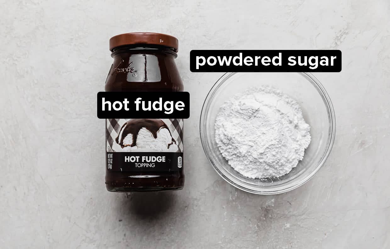 A jar of hot fudge sauce and a bowl of powdered sugar on a gray background.