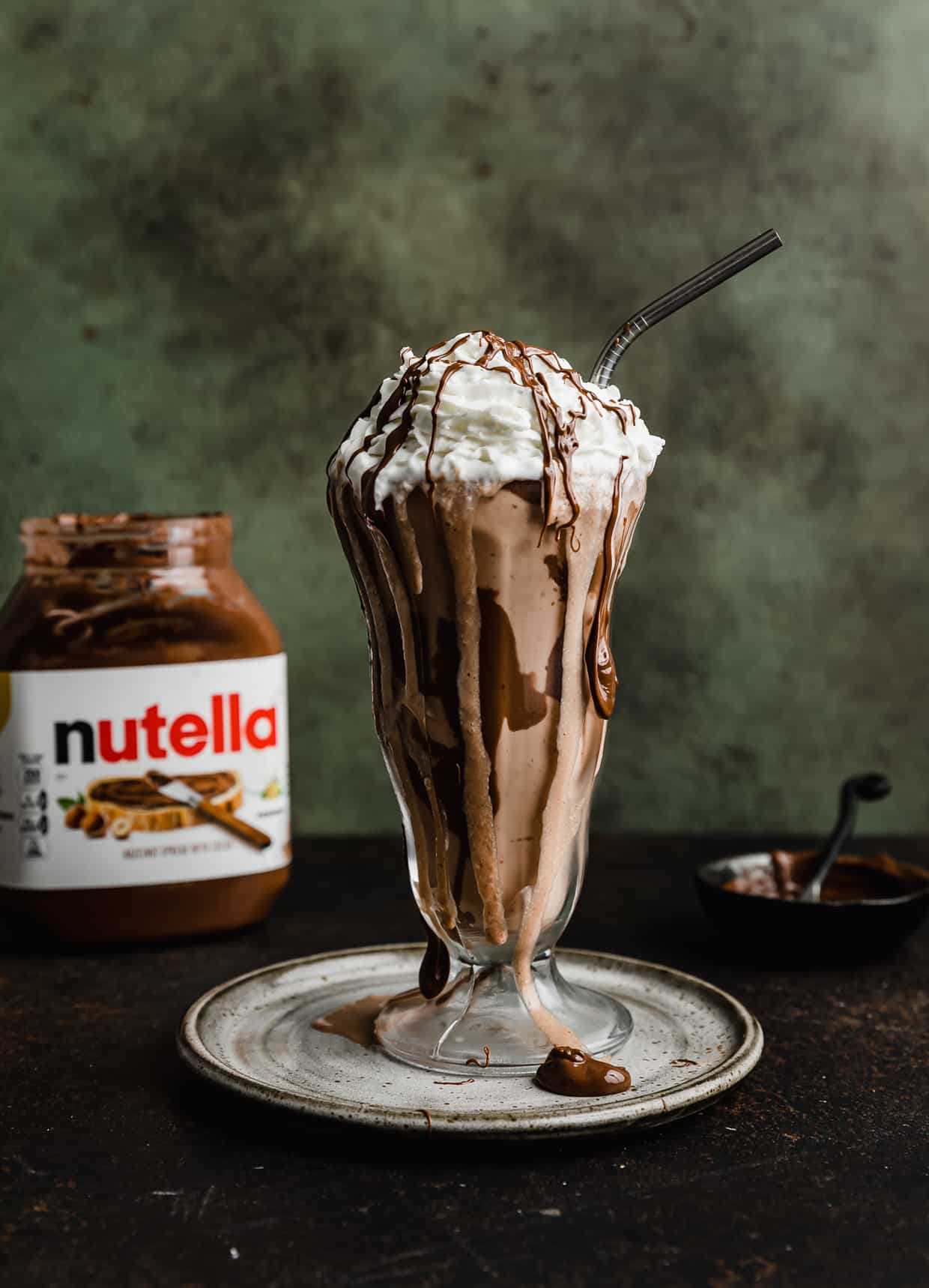 A glass milkshake cup with a Nutella Milkshake in it, topped with whipped topping and drizzled Nutella in front of a dark green background.